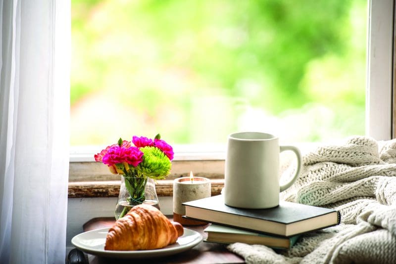 A mug sits atop a stack of books next to a croissant, flowes, and lit candles next to a bright window representing a hygge style afternoon.