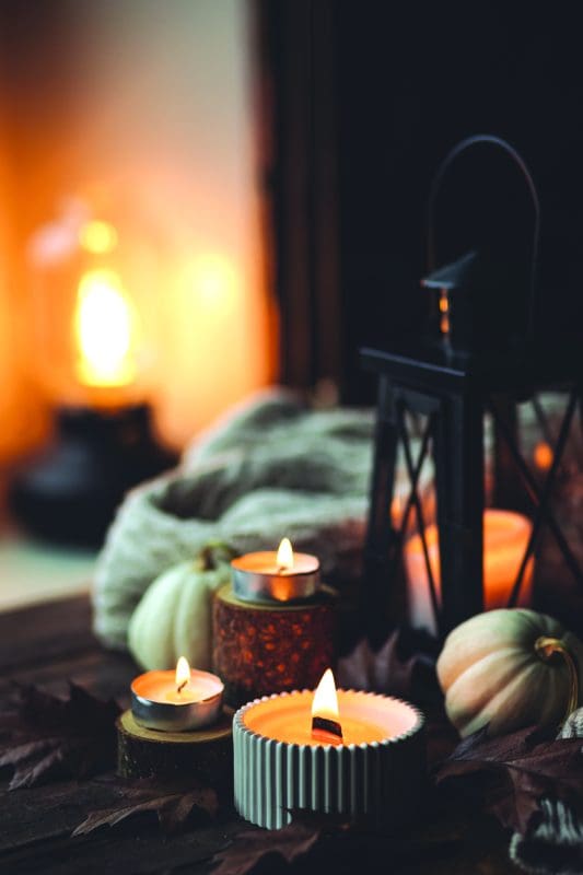 Several lit candles and a lantern bathed in warm light, surrounded by fall leaves, pumpkins, and blankets, exude the hygge principal of light.