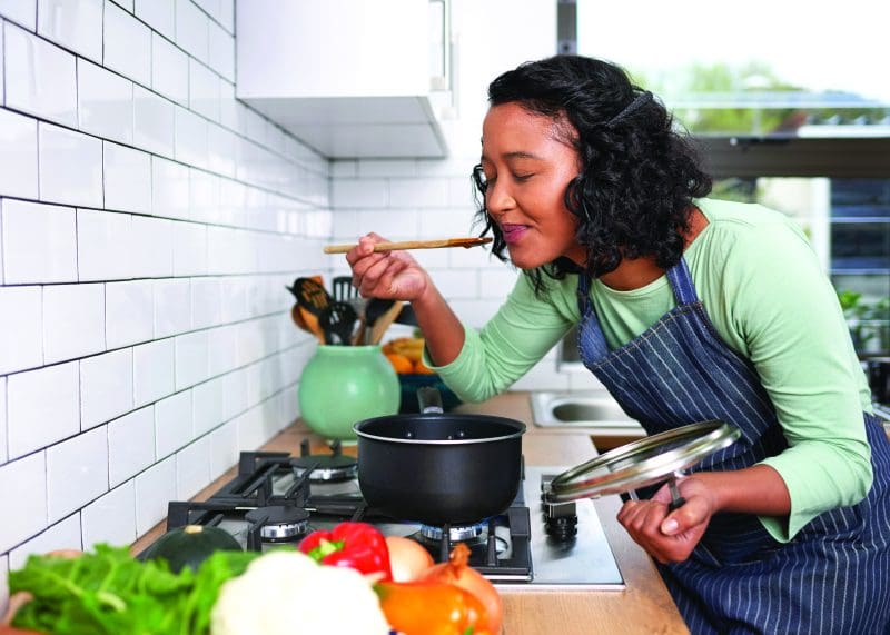 A woman in an apron in a brightly lit kitchen tasted a soup on the stove, practicing the slow cooking tenet of hygge.