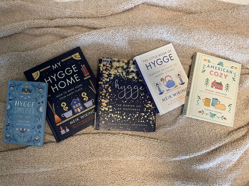 Four books about hygge spread out on a cozy blanket. The books in order from left to right are Hygge Simplified, My Hygge Home, Hygge (the Danish Art of Happiness), The Little Book of Hygge, and American Cozy.